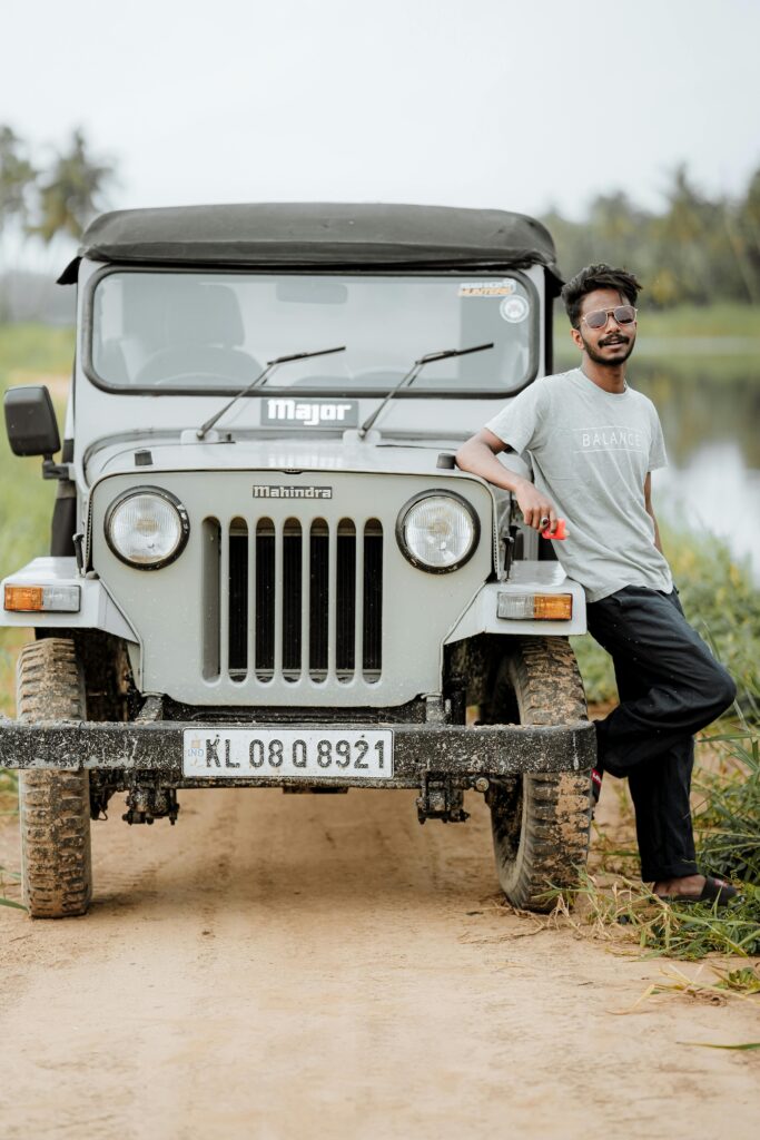 Young man in sunglasses leans on a rugged off-road vehicle on a dirt path by a water body.
